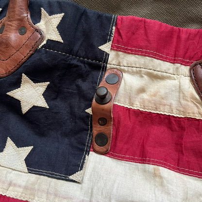 Ralph Lauren Denim &amp; Supply 1990s USA Flag Shoulder Bag is a large shoulder bag constructed from a mix of leather and cotton canvas. One side features a faded USA flag made from stitched canvas patches, while the reverse side is khaki canvas. The bag includes leather handles and a leather strap closure, an interior main compartment with additional smaller pockets, and a Denim &amp; Supply tag.
