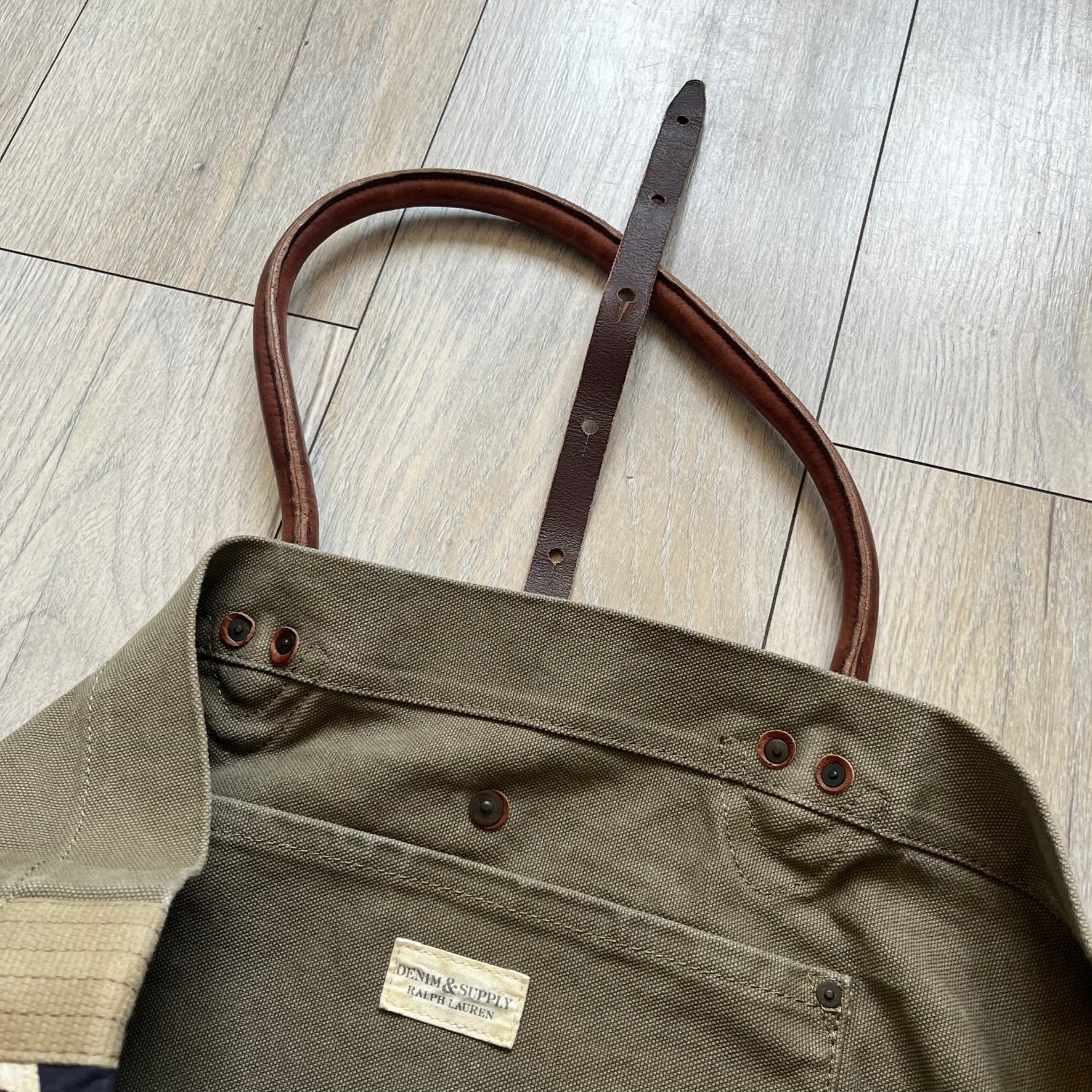 Ralph Lauren Denim &amp; Supply 1990s USA Flag Shoulder Bag is a large shoulder bag constructed from a mix of leather and cotton canvas. One side features a faded USA flag made from stitched canvas patches, while the reverse side is khaki canvas. The bag includes leather handles and a leather strap closure, an interior main compartment with additional smaller pockets, and a Denim &amp; Supply tag.
