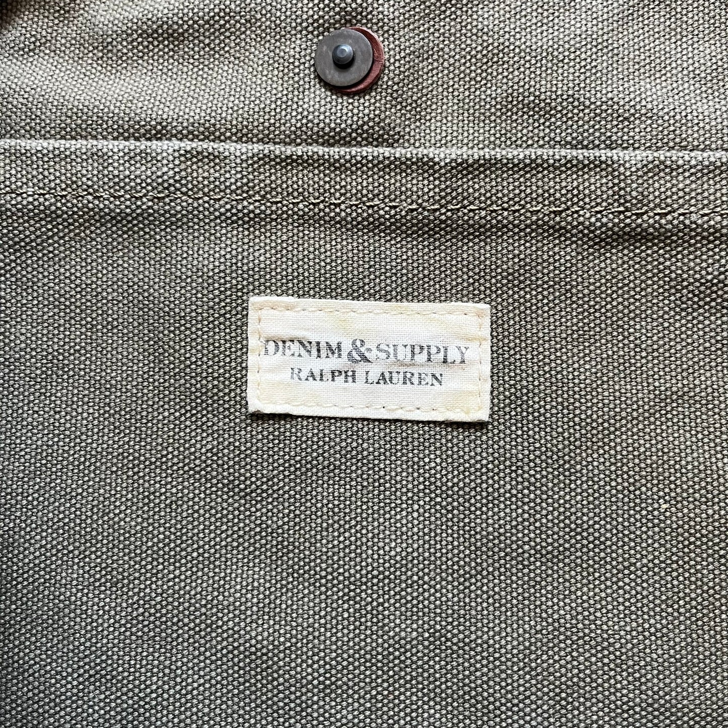 Ralph Lauren Denim &amp; Supply 1990s USA Flag Shoulder Bag is a large shoulder bag constructed from a mix of leather and cotton canvas. One side features a faded USA flag made from stitched canvas patches, while the reverse side is khaki canvas. The bag includes leather handles and a leather strap closure, an interior main compartment with additional smaller pockets, and a Denim &amp; Supply tag.
