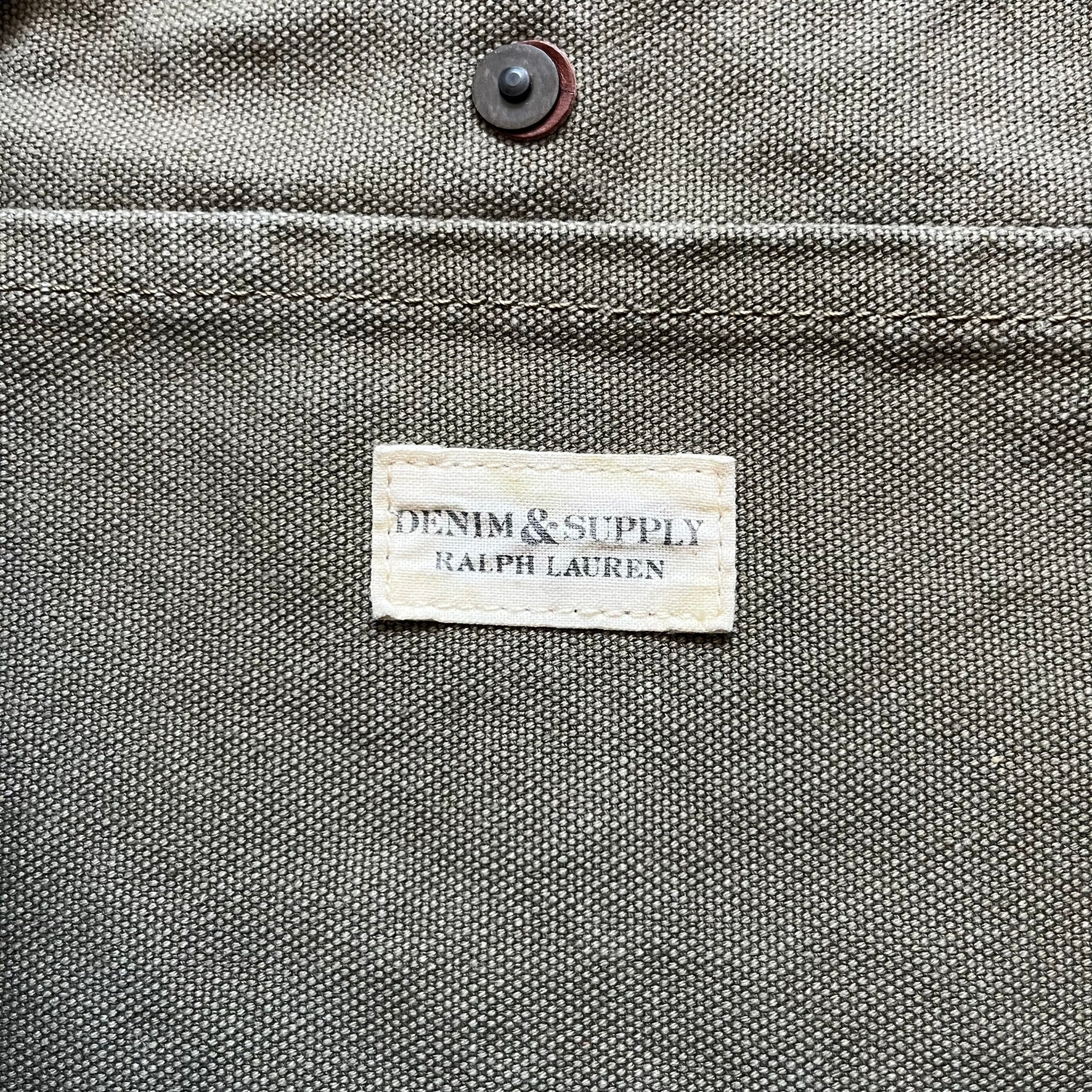 Ralph Lauren Denim &amp; Supply 1990s USA Flag Shoulder Bag is a large shoulder bag constructed from a mix of leather and cotton canvas. One side features a faded USA flag made from stitched canvas patches, while the reverse side is khaki canvas. The bag includes leather handles and a leather strap closure, an interior main compartment with additional smaller pockets, and a Denim &amp; Supply tag.
