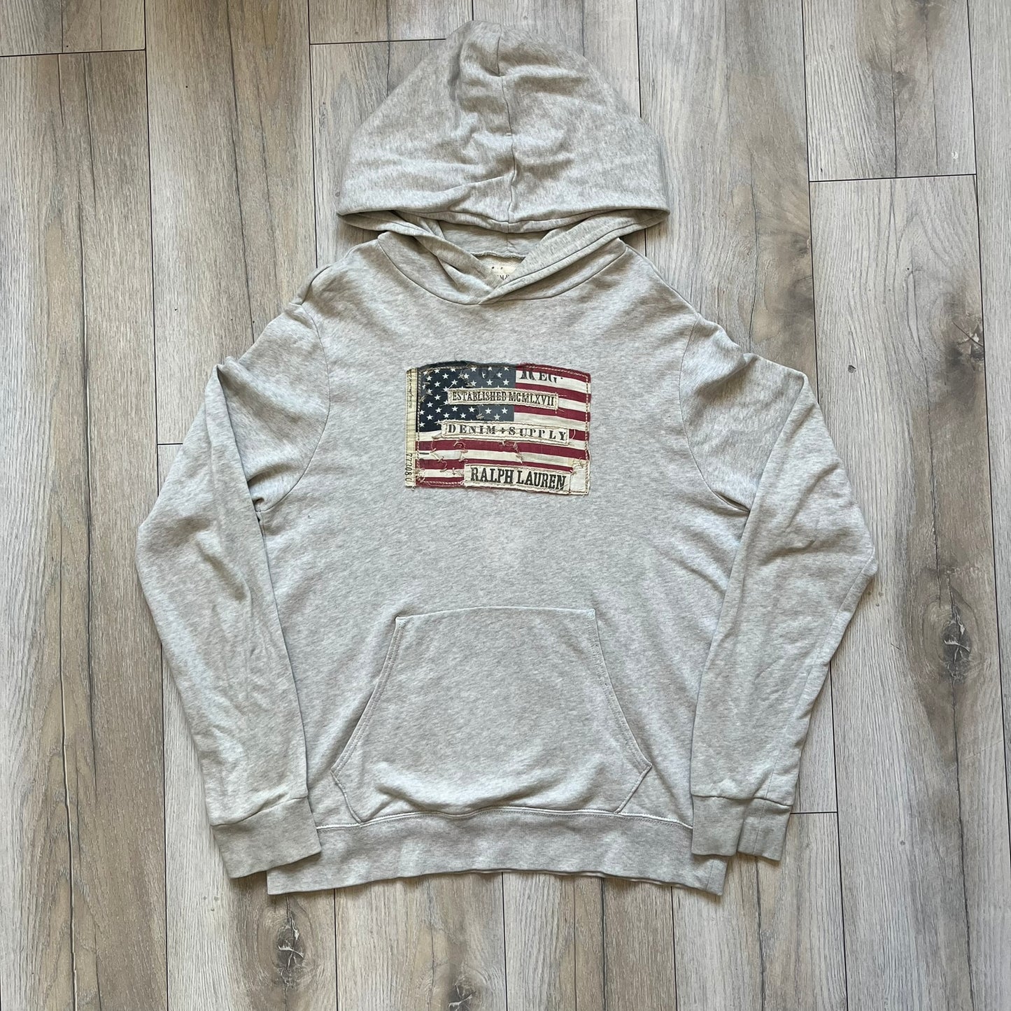 Ralph Lauren Denim & Supply USA Flag Hoodie is a grey pullover hoodie featuring a large USA flag patch on the chest constructed from multiple stitched elements. The flag includes “Denim & Supply,” “Ralph Lauren,” and “Established MCMLXVII” details, with a distressed, multi-panel construction. The hoodie has a Denim & Supply neck tag, large hood, and a standard front kangaroo pocket.