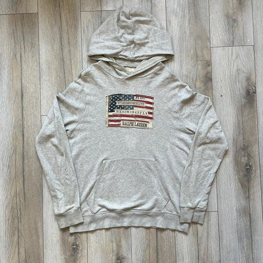 Ralph Lauren Denim & Supply USA Flag Hoodie is a grey pullover hoodie featuring a large USA flag patch on the chest constructed from multiple stitched elements. The flag includes “Denim & Supply,” “Ralph Lauren,” and “Established MCMLXVII” details, with a distressed, multi-panel construction. The hoodie has a Denim & Supply neck tag, large hood, and a standard front kangaroo pocket.