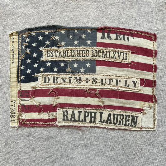 Ralph Lauren Denim & Supply USA Flag Hoodie is a grey pullover hoodie featuring a large USA flag patch on the chest constructed from multiple stitched elements. The flag includes “Denim & Supply,” “Ralph Lauren,” and “Established MCMLXVII” details, with a distressed, multi-panel construction. The hoodie has a Denim & Supply neck tag, large hood, and a standard front kangaroo pocket.