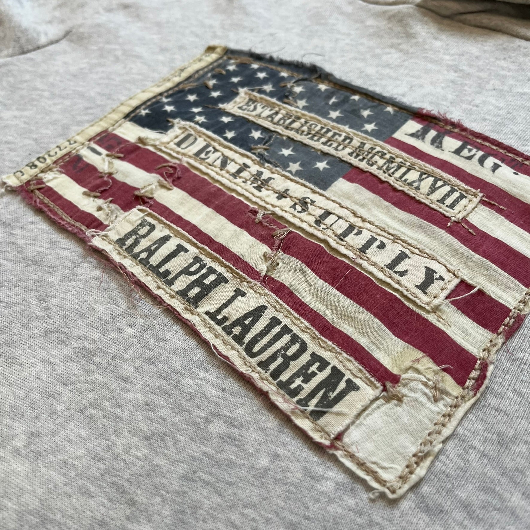 Ralph Lauren Denim & Supply USA Flag Hoodie is a grey pullover hoodie featuring a large USA flag patch on the chest constructed from multiple stitched elements. The flag includes “Denim & Supply,” “Ralph Lauren,” and “Established MCMLXVII” details, with a distressed, multi-panel construction. The hoodie has a Denim & Supply neck tag, large hood, and a standard front kangaroo pocket.