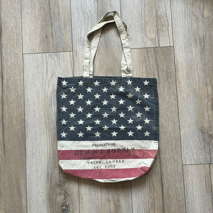 Ralph Lauren Denim &amp; Supply USA Flag Tote Bag is a fabric tote featuring an all-over faded effect. Both sides display a USA flag print with white hand graphics integrated into the design. The bottom includes printed branding, with “Property of Denim &amp; Supply” on one side and “Denim &amp; Supply Ralph Lauren” on the other.
