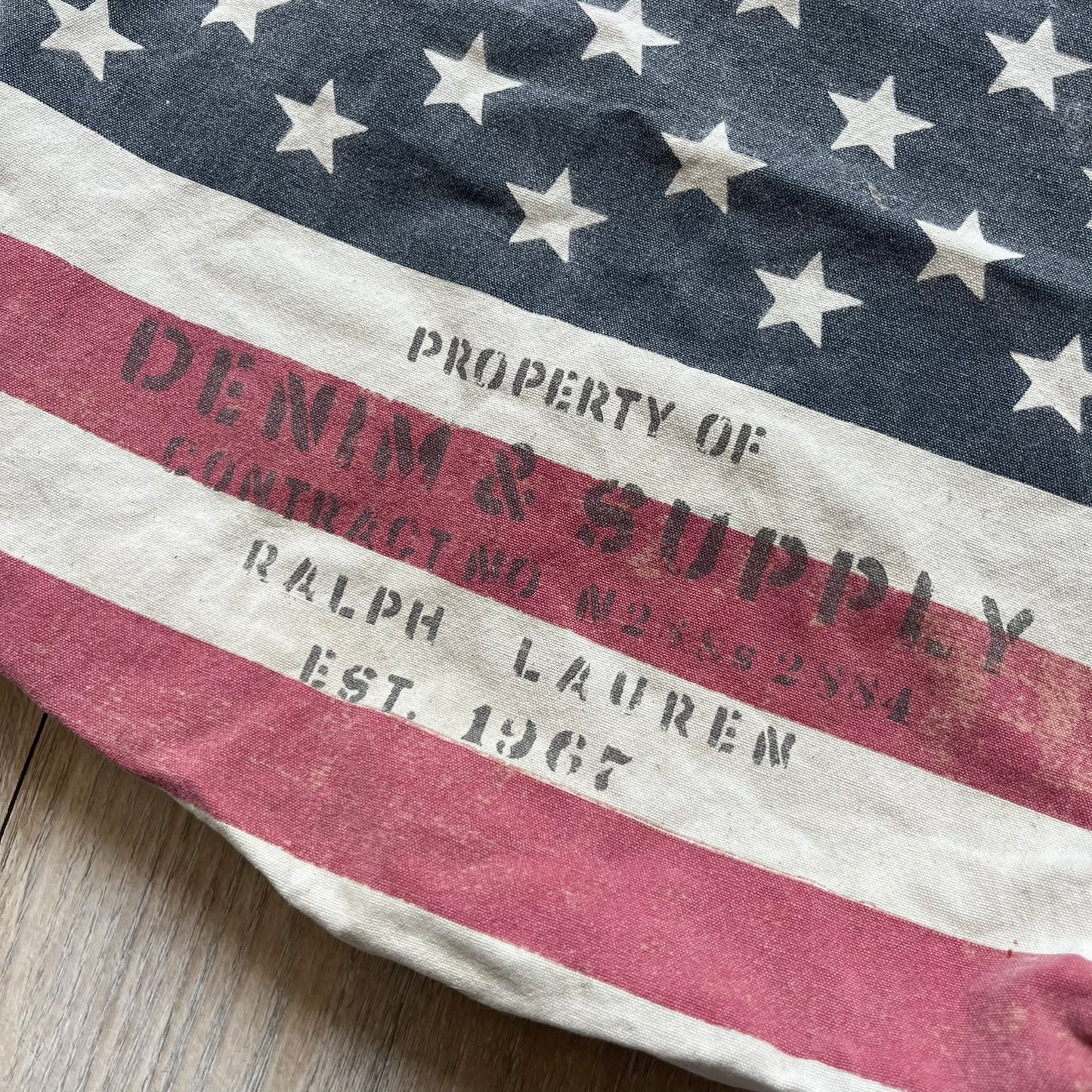 Ralph Lauren Denim &amp; Supply USA Flag Tote Bag is a fabric tote featuring an all-over faded effect. Both sides display a USA flag print with white hand graphics integrated into the design. The bottom includes printed branding, with “Property of Denim &amp; Supply” on one side and “Denim &amp; Supply Ralph Lauren” on the other.
