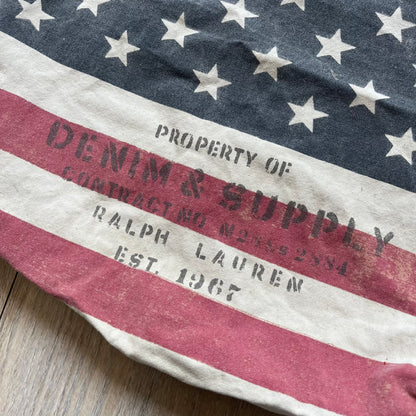 Ralph Lauren Denim &amp; Supply USA Flag Tote Bag is a fabric tote featuring an all-over faded effect. Both sides display a USA flag print with white hand graphics integrated into the design. The bottom includes printed branding, with “Property of Denim &amp; Supply” on one side and “Denim &amp; Supply Ralph Lauren” on the other.
