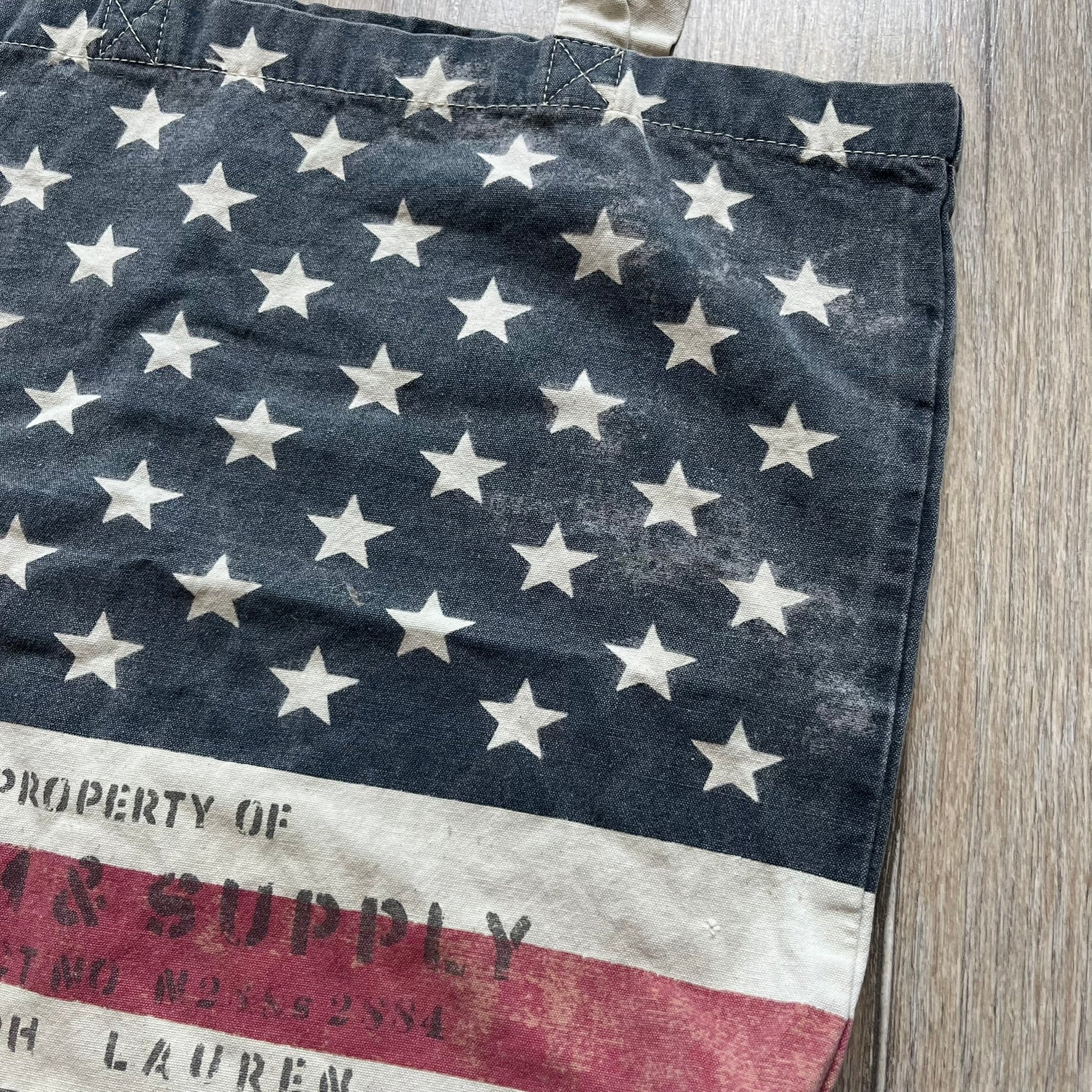 Ralph Lauren Denim &amp; Supply USA Flag Tote Bag is a fabric tote featuring an all-over faded effect. Both sides display a USA flag print with white hand graphics integrated into the design. The bottom includes printed branding, with “Property of Denim &amp; Supply” on one side and “Denim &amp; Supply Ralph Lauren” on the other.
