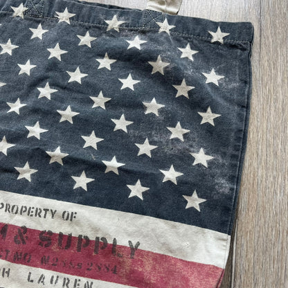 Ralph Lauren Denim &amp; Supply USA Flag Tote Bag is a fabric tote featuring an all-over faded effect. Both sides display a USA flag print with white hand graphics integrated into the design. The bottom includes printed branding, with “Property of Denim &amp; Supply” on one side and “Denim &amp; Supply Ralph Lauren” on the other.

