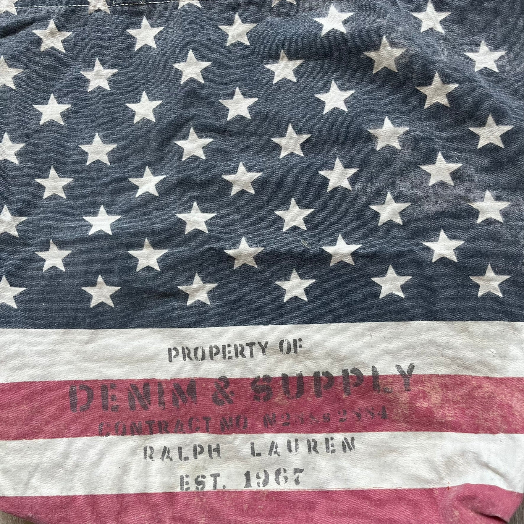 Ralph Lauren Denim &amp; Supply USA Flag Tote Bag is a fabric tote featuring an all-over faded effect. Both sides display a USA flag print with white hand graphics integrated into the design. The bottom includes printed branding, with “Property of Denim &amp; Supply” on one side and “Denim &amp; Supply Ralph Lauren” on the other.
