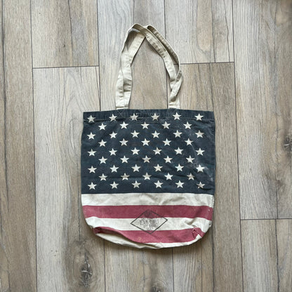 Ralph Lauren Denim &amp; Supply USA Flag Tote Bag is a fabric tote featuring an all-over faded effect. Both sides display a USA flag print with white hand graphics integrated into the design. The bottom includes printed branding, with “Property of Denim &amp; Supply” on one side and “Denim &amp; Supply Ralph Lauren” on the other.
