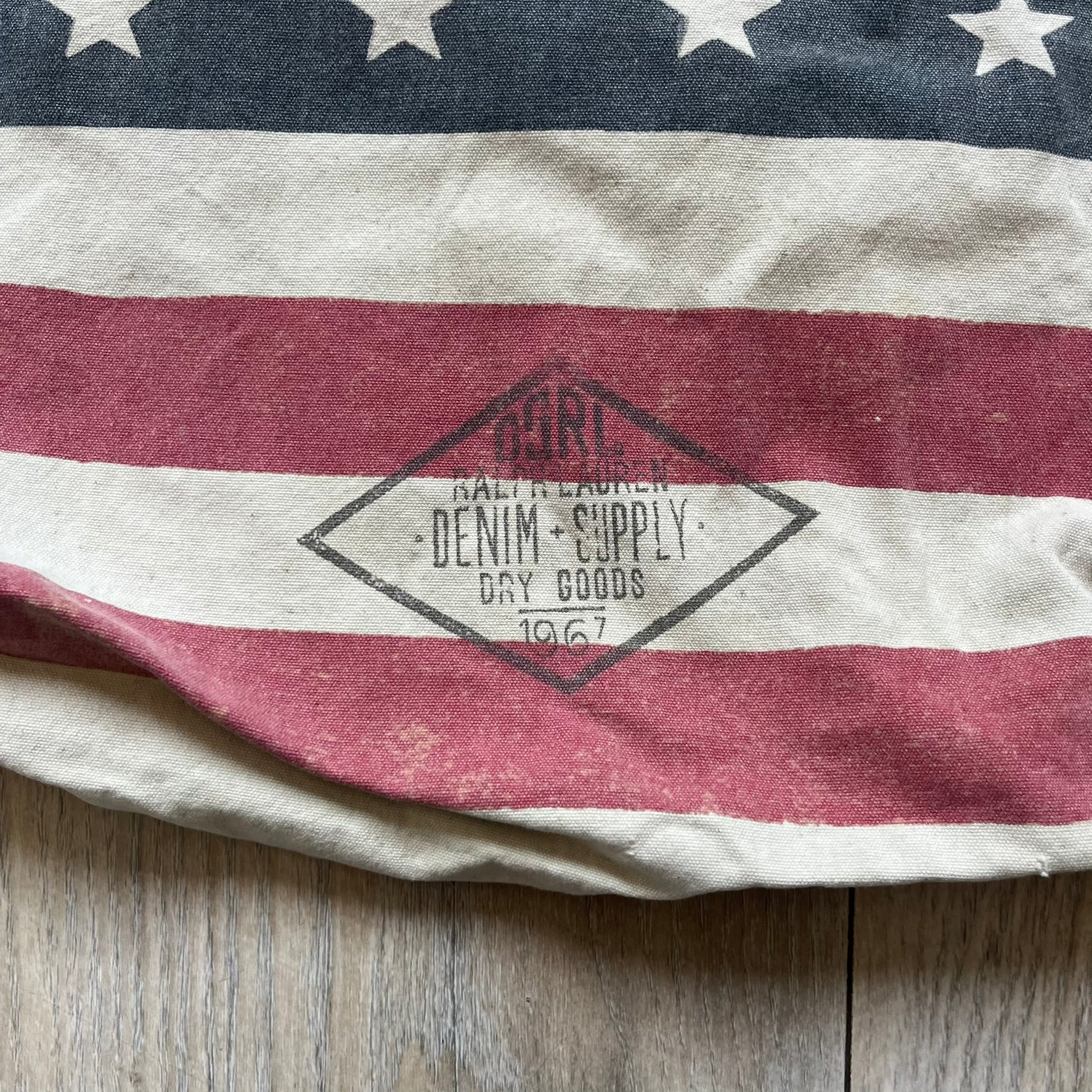 Ralph Lauren Denim &amp; Supply USA Flag Tote Bag is a fabric tote featuring an all-over faded effect. Both sides display a USA flag print with white hand graphics integrated into the design. The bottom includes printed branding, with “Property of Denim &amp; Supply” on one side and “Denim &amp; Supply Ralph Lauren” on the other.
