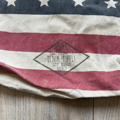Ralph Lauren Denim &amp; Supply USA Flag Tote Bag is a fabric tote featuring an all-over faded effect. Both sides display a USA flag print with white hand graphics integrated into the design. The bottom includes printed branding, with “Property of Denim &amp; Supply” on one side and “Denim &amp; Supply Ralph Lauren” on the other.
