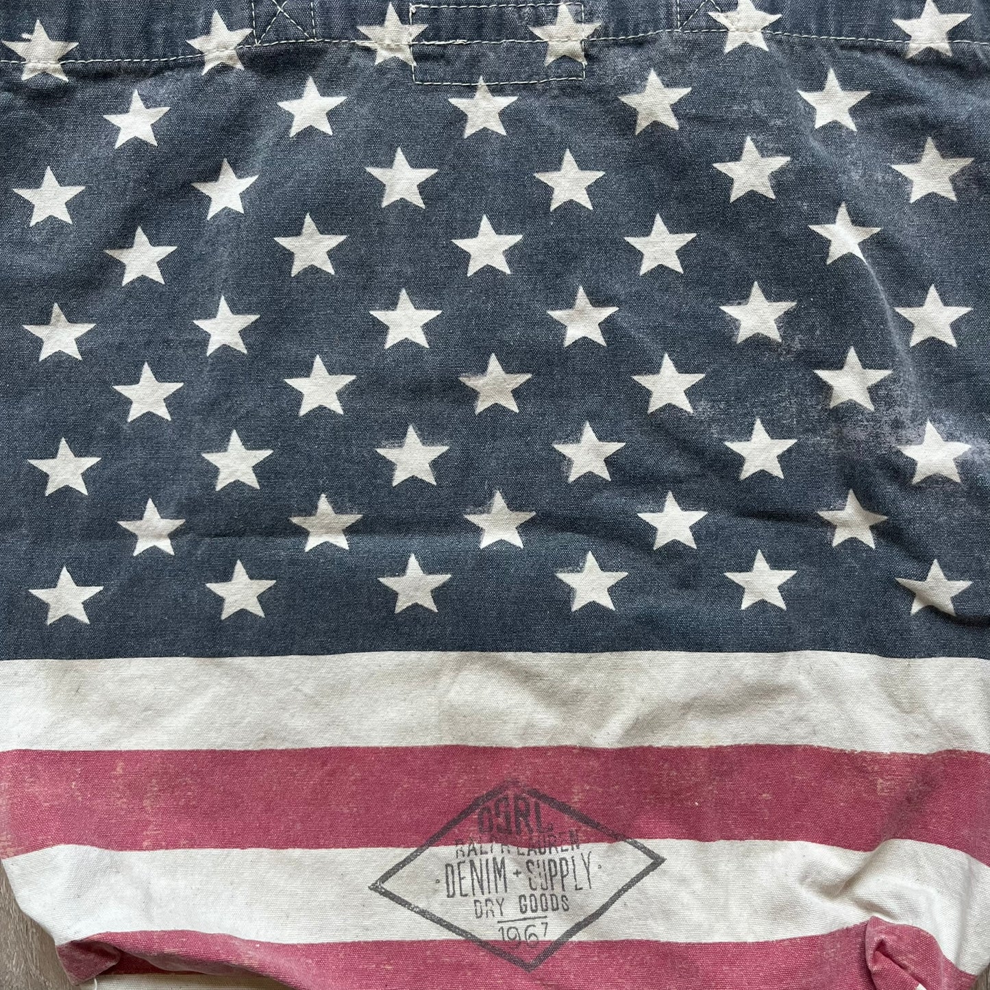 Ralph Lauren Denim &amp; Supply USA Flag Tote Bag is a fabric tote featuring an all-over faded effect. Both sides display a USA flag print with white hand graphics integrated into the design. The bottom includes printed branding, with “Property of Denim &amp; Supply” on one side and “Denim &amp; Supply Ralph Lauren” on the other.
