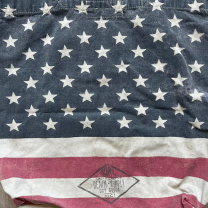 Ralph Lauren Denim &amp; Supply USA Flag Tote Bag is a fabric tote featuring an all-over faded effect. Both sides display a USA flag print with white hand graphics integrated into the design. The bottom includes printed branding, with “Property of Denim &amp; Supply” on one side and “Denim &amp; Supply Ralph Lauren” on the other.
