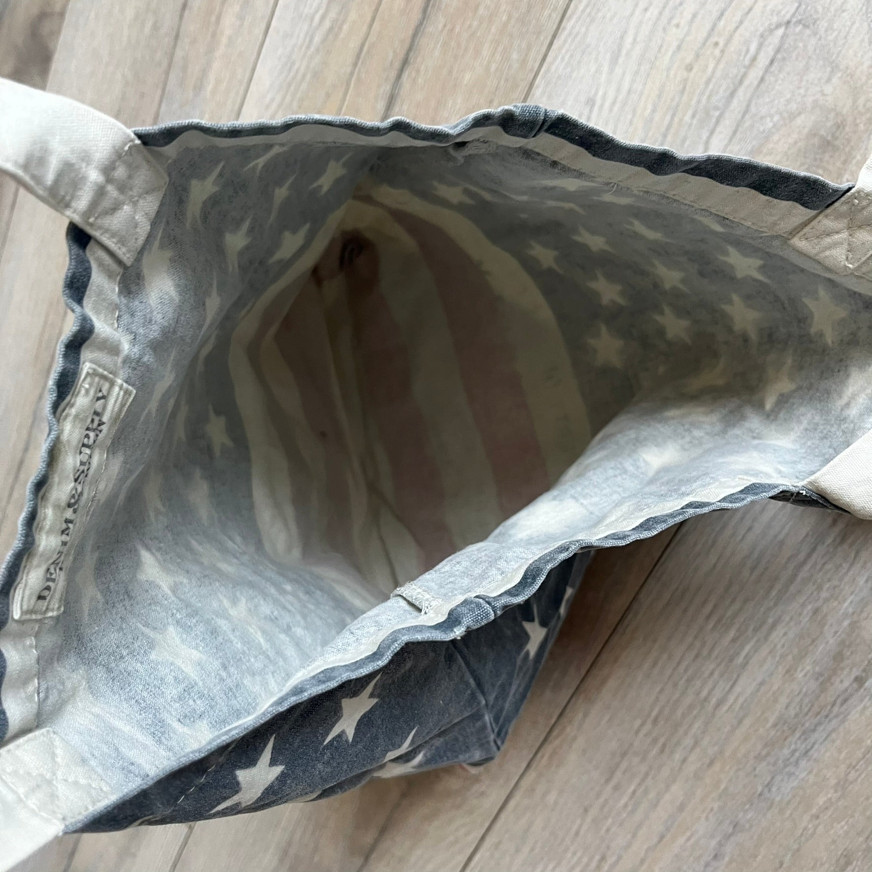 Ralph Lauren Denim &amp; Supply USA Flag Tote Bag is a fabric tote featuring an all-over faded effect. Both sides display a USA flag print with white hand graphics integrated into the design. The bottom includes printed branding, with “Property of Denim &amp; Supply” on one side and “Denim &amp; Supply Ralph Lauren” on the other.
