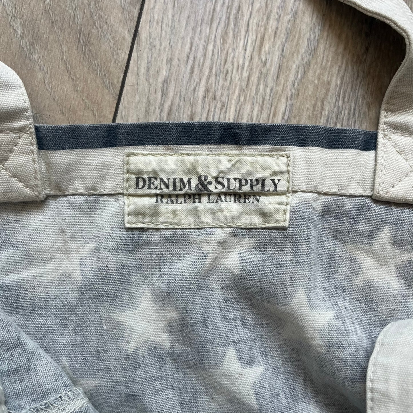 Ralph Lauren Denim &amp; Supply USA Flag Tote Bag is a fabric tote featuring an all-over faded effect. Both sides display a USA flag print with white hand graphics integrated into the design. The bottom includes printed branding, with “Property of Denim &amp; Supply” on one side and “Denim &amp; Supply Ralph Lauren” on the other.
