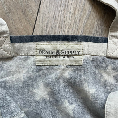 Ralph Lauren Denim &amp; Supply USA Flag Tote Bag is a fabric tote featuring an all-over faded effect. Both sides display a USA flag print with white hand graphics integrated into the design. The bottom includes printed branding, with “Property of Denim &amp; Supply” on one side and “Denim &amp; Supply Ralph Lauren” on the other.
