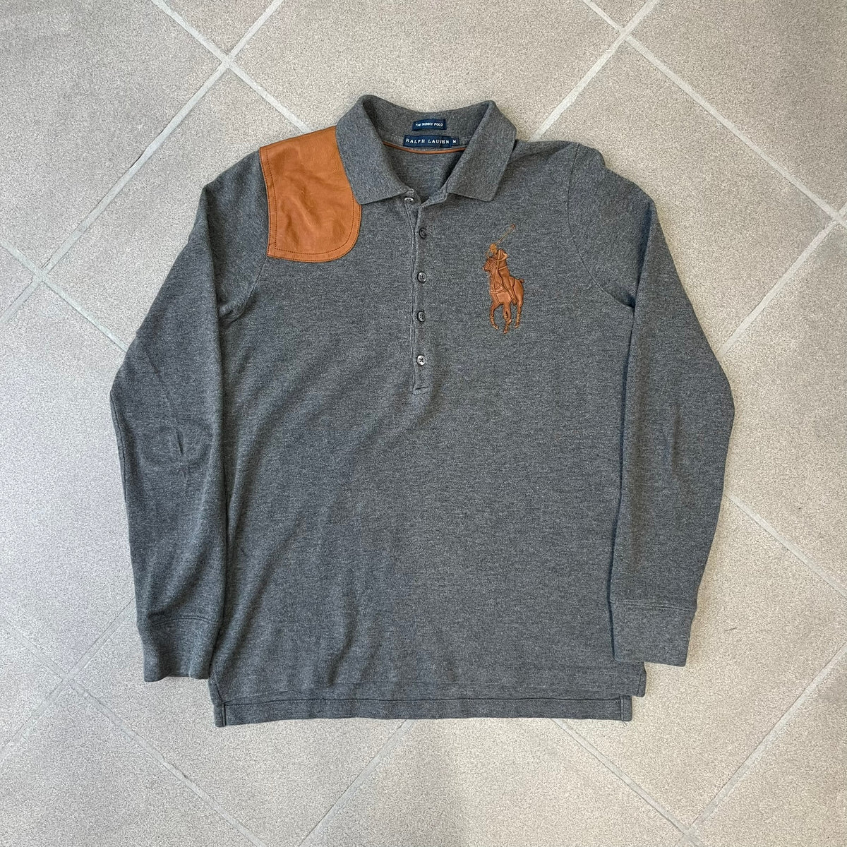Ralph Lauren Leather Patch Gray Polo Longsleeve features a grey 100% cotton longsleeve polo with a brown leather patch on the right shoulder. The left chest has a raised 3D leather Polo logo, while the shirt has a button closure and is tagged “The Skinny Polo.”