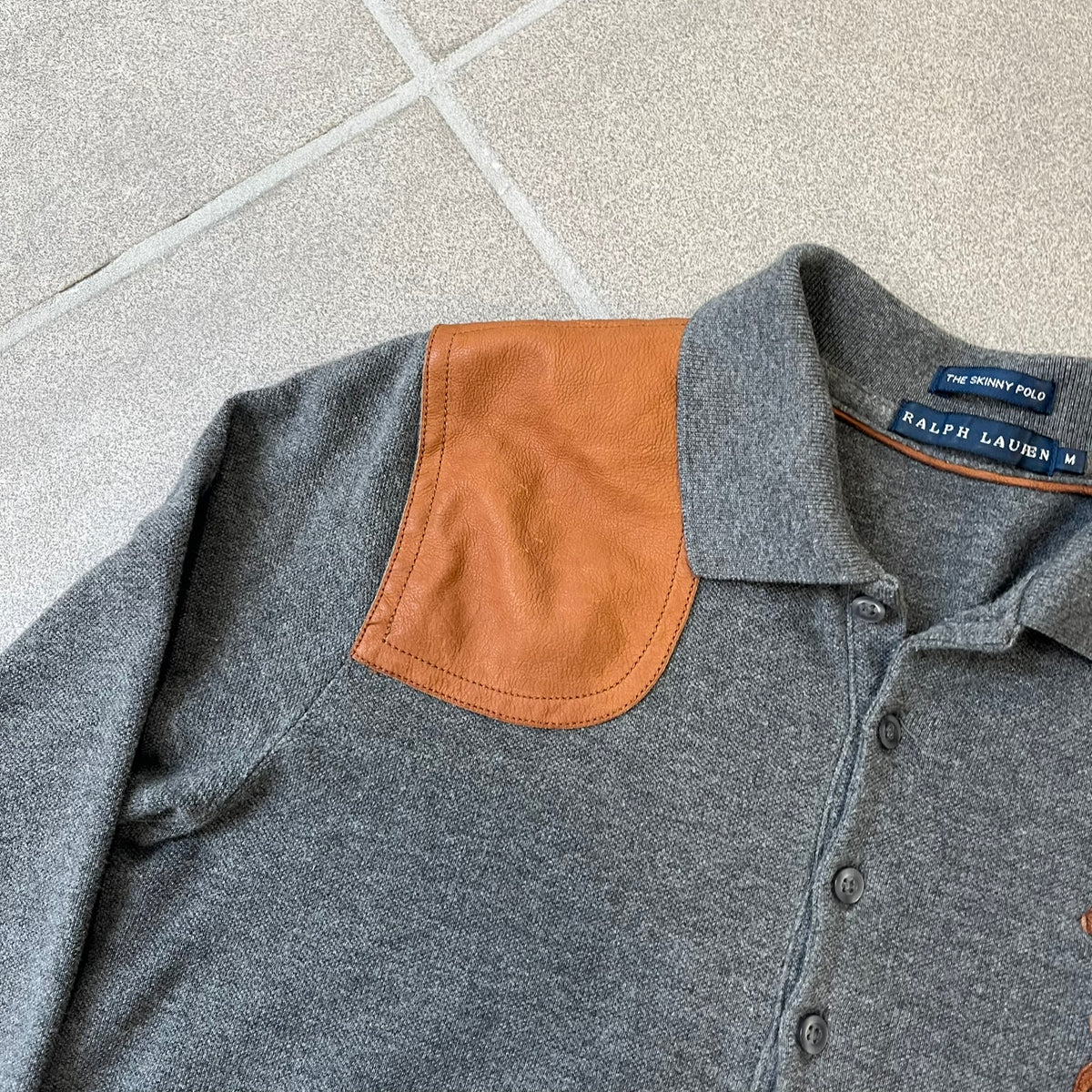 Ralph Lauren Leather Patch Gray Polo Longsleeve features a grey 100% cotton longsleeve polo with a brown leather patch on the right shoulder. The left chest has a raised 3D leather Polo logo, while the shirt has a button closure and is tagged “The Skinny Polo.”