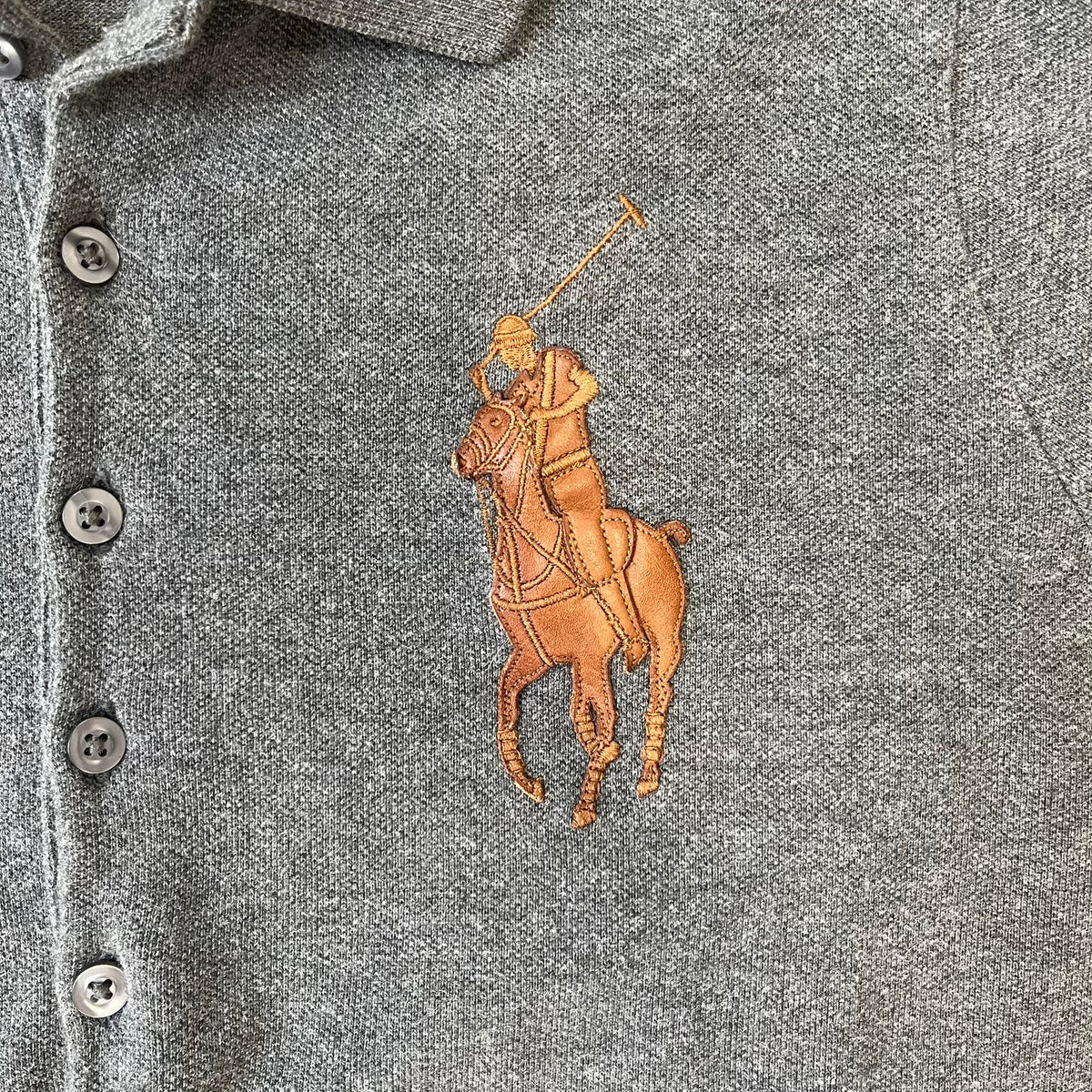 Ralph Lauren Leather Patch Gray Polo Longsleeve features a grey 100% cotton longsleeve polo with a brown leather patch on the right shoulder. The left chest has a raised 3D leather Polo logo, while the shirt has a button closure and is tagged “The Skinny Polo.”