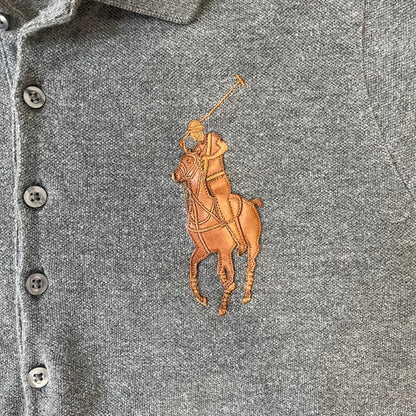 Ralph Lauren Leather Patch Gray Polo Longsleeve features a grey 100% cotton longsleeve polo with a brown leather patch on the right shoulder. The left chest has a raised 3D leather Polo logo, while the shirt has a button closure and is tagged “The Skinny Polo.”