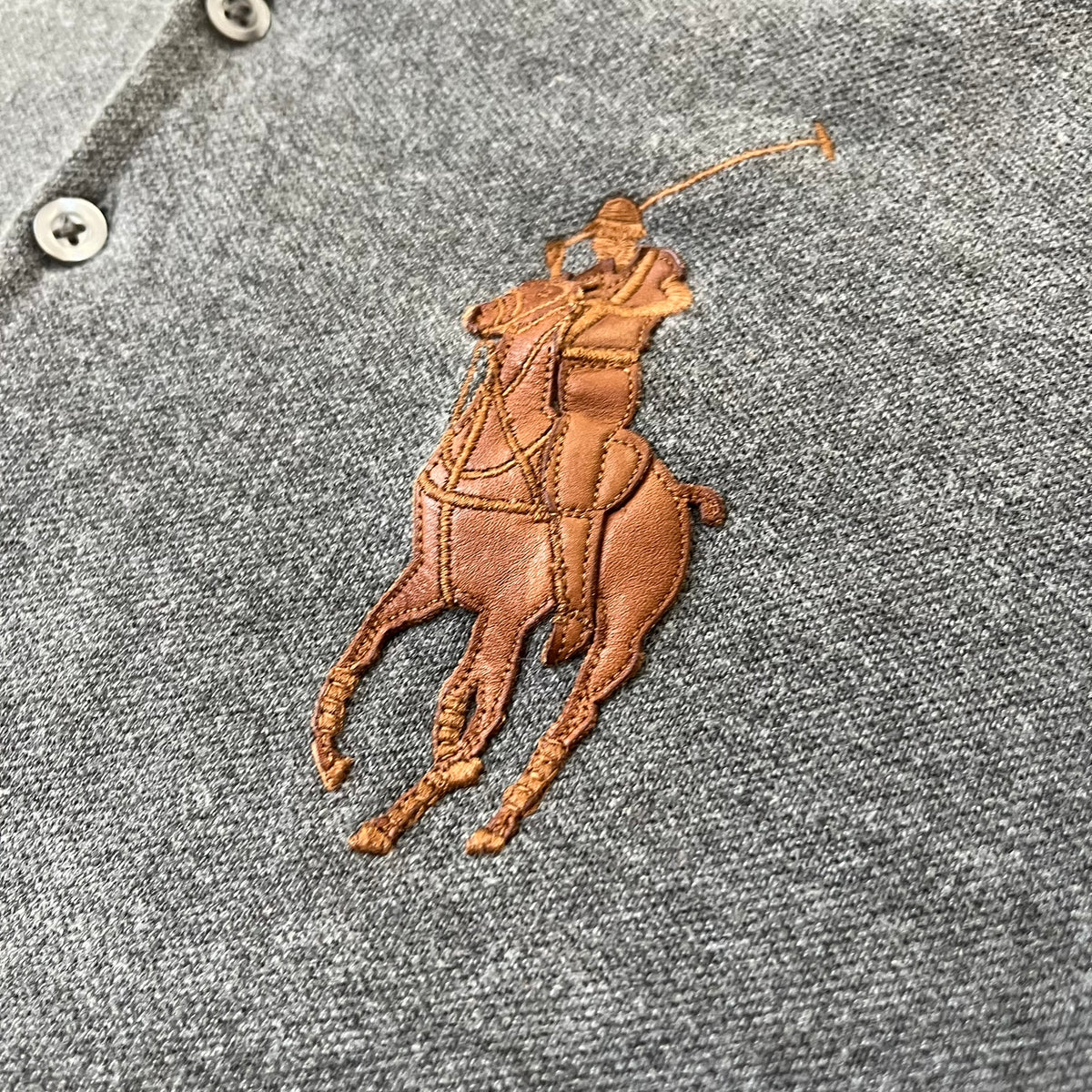 Ralph Lauren Leather Patch Gray Polo Longsleeve features a grey 100% cotton longsleeve polo with a brown leather patch on the right shoulder. The left chest has a raised 3D leather Polo logo, while the shirt has a button closure and is tagged “The Skinny Polo.”