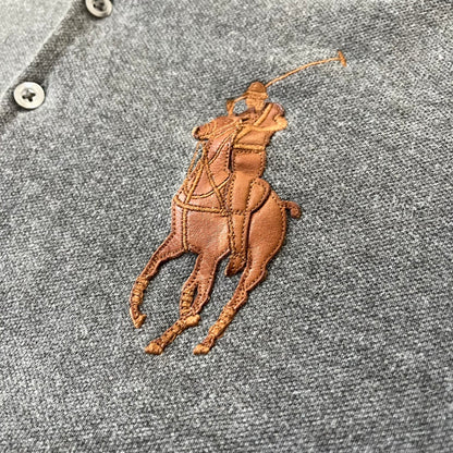 Ralph Lauren Leather Patch Gray Polo Longsleeve features a grey 100% cotton longsleeve polo with a brown leather patch on the right shoulder. The left chest has a raised 3D leather Polo logo, while the shirt has a button closure and is tagged “The Skinny Polo.”