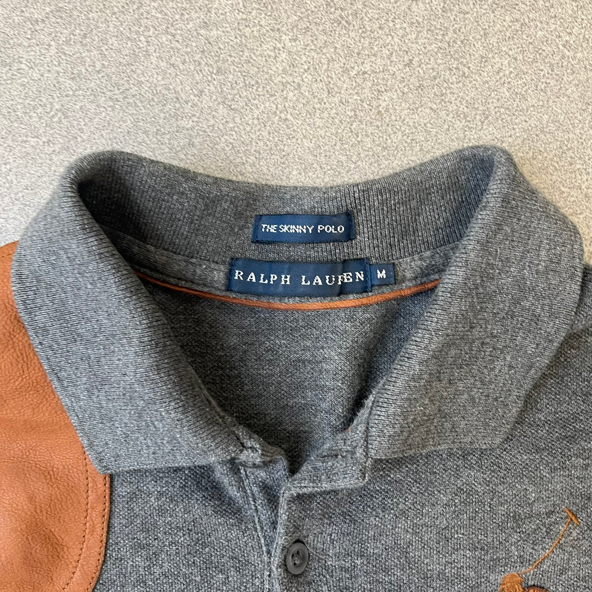 Ralph Lauren Leather Patch Gray Polo Longsleeve features a grey 100% cotton longsleeve polo with a brown leather patch on the right shoulder. The left chest has a raised 3D leather Polo logo, while the shirt has a button closure and is tagged “The Skinny Polo.”