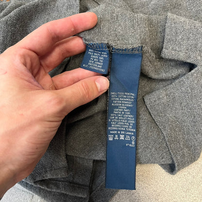 Ralph Lauren Leather Patch Gray Polo Longsleeve features a grey 100% cotton longsleeve polo with a brown leather patch on the right shoulder. The left chest has a raised 3D leather Polo logo, while the shirt has a button closure and is tagged “The Skinny Polo.”