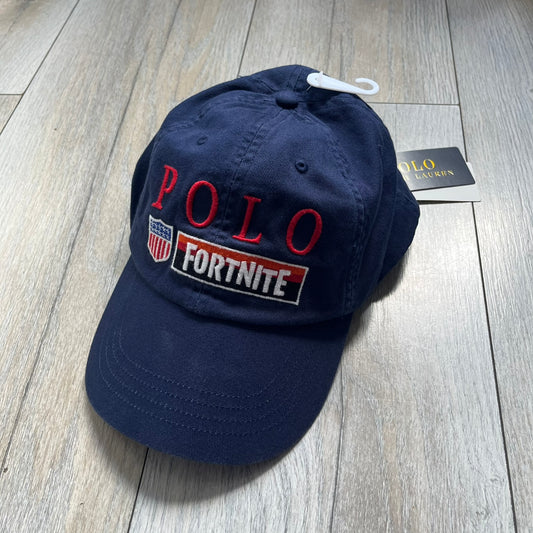 Ralph Lauren x Fortnite Navy Logo Baseball Cap is a navy cotton cap featuring bold embroidered branding on the front, including red Polo Ralph Lauren text and a Fortnite logo patch combined with a USA Ralph Lauren crest. The back includes a white embroidered Ralph Lauren logo and an adjustable strap for a custom fit.
