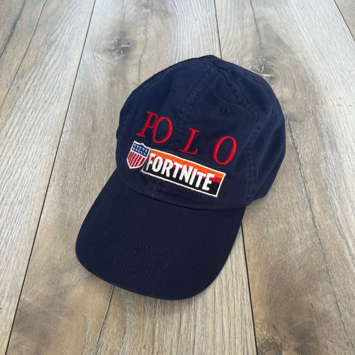 Ralph Lauren x Fortnite Navy Logo Baseball Cap is a navy cotton cap featuring bold embroidered branding on the front, including red Polo Ralph Lauren text and a Fortnite logo patch combined with a USA Ralph Lauren crest. The back includes a white embroidered Ralph Lauren logo and an adjustable strap for a custom fit.