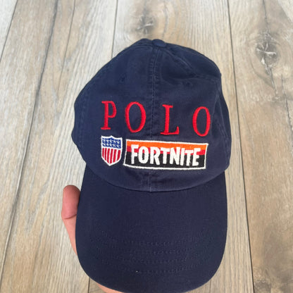 Ralph Lauren x Fortnite Navy Logo Baseball Cap is a navy cotton cap featuring bold embroidered branding on the front, including red Polo Ralph Lauren text and a Fortnite logo patch combined with a USA Ralph Lauren crest. The back includes a white embroidered Ralph Lauren logo and an adjustable strap for a custom fit.