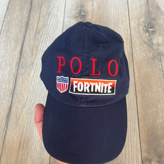 Ralph Lauren x Fortnite Navy Logo Baseball Cap is a navy cotton cap featuring bold embroidered branding on the front, including red Polo Ralph Lauren text and a Fortnite logo patch combined with a USA Ralph Lauren crest. The back includes a white embroidered Ralph Lauren logo and an adjustable strap for a custom fit.