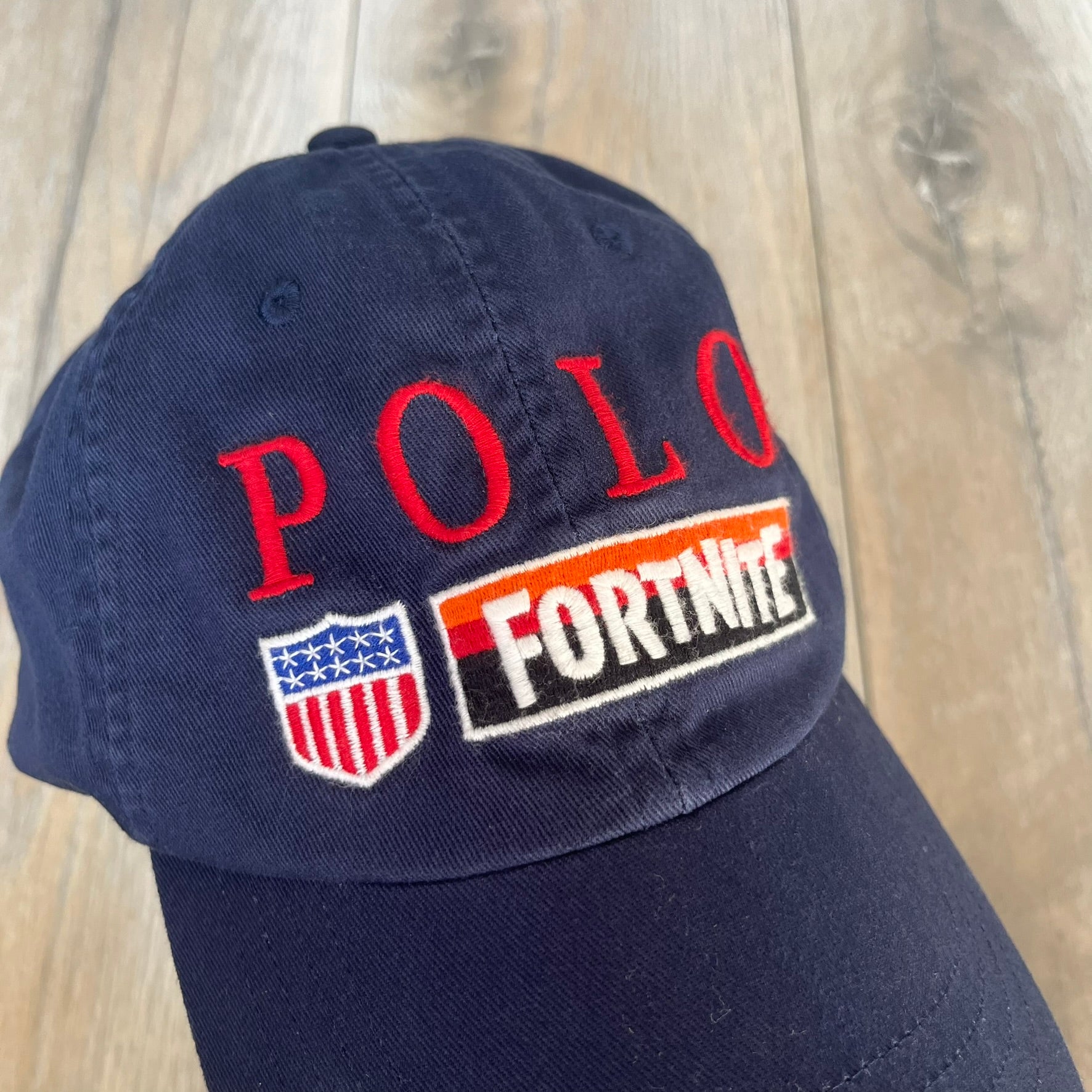 Ralph Lauren x Fortnite Navy Logo Baseball Cap is a navy cotton cap featuring bold embroidered branding on the front, including red Polo Ralph Lauren text and a Fortnite logo patch combined with a USA Ralph Lauren crest. The back includes a white embroidered Ralph Lauren logo and an adjustable strap for a custom fit.