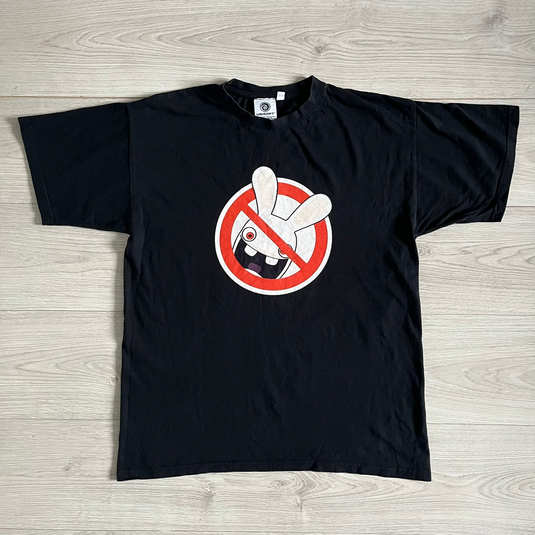 Raving Rabbids - known as “Lapins Crétins” in France - 2010 Tee in black, features a white Rabbid head inside a red circle with a slash, similar to a forbidden symbol. Officially licensed by Ubisoft, it comes with branding tags for collector and fan authenticity.&nbsp;