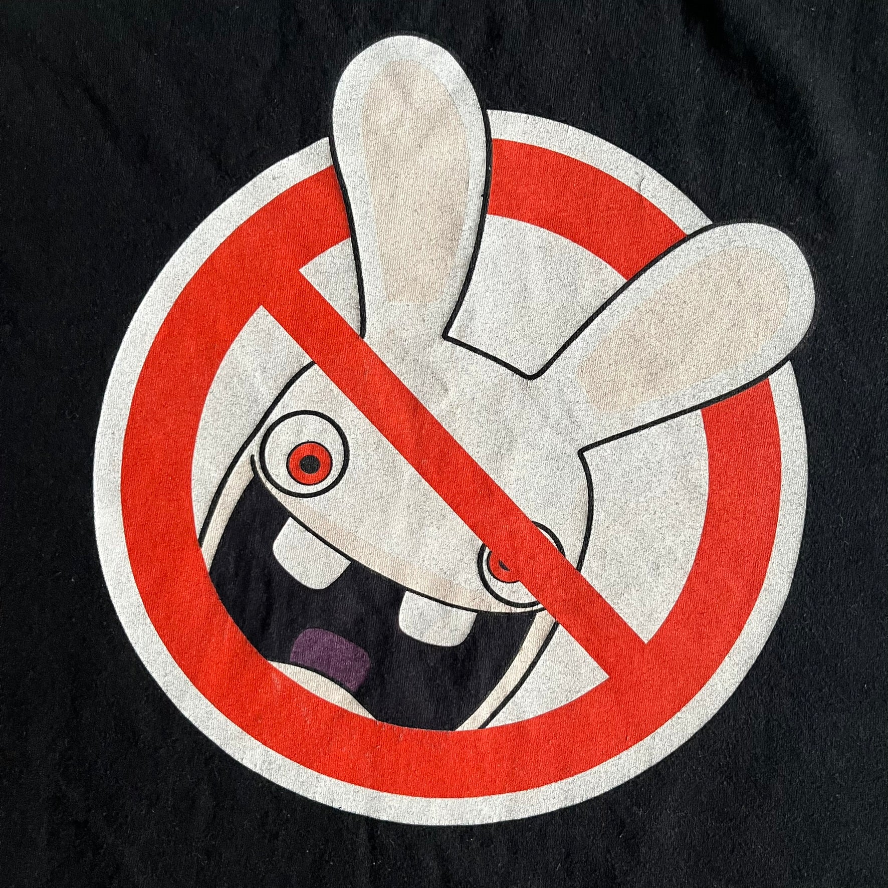 Raving Rabbids - known as “Lapins Crétins” in France - 2010 Tee in black, features a white Rabbid head inside a red circle with a slash, similar to a forbidden symbol. Officially licensed by Ubisoft, it comes with branding tags for collector and fan authenticity.&nbsp;
