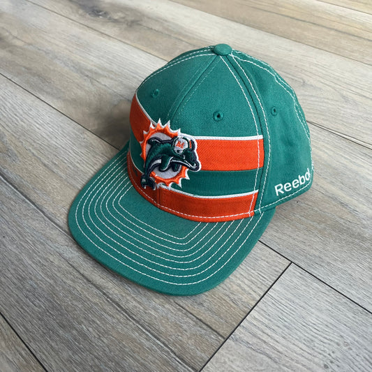 Reebok NFL Miami Dolphins Onfield Snapback Cap is a turquoise snapback featuring embroidered Miami Dolphins branding on the front, framed by two bold orange stripes positioned at the top and bottom of the front panel. The visor includes white stitching on the top surface and an orange underside for contrast. The side is detailed with a white Reebok logo, while the back features NFL Equipment and Onfield branding.