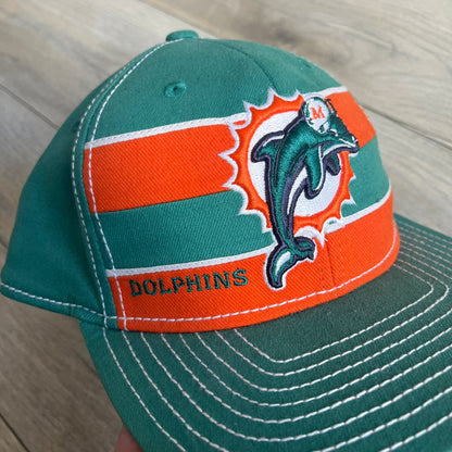 Reebok NFL Miami Dolphins Onfield Snapback Cap is a turquoise snapback featuring embroidered Miami Dolphins branding on the front, framed by two bold orange stripes positioned at the top and bottom of the front panel. The visor includes white stitching on the top surface and an orange underside for contrast. The side is detailed with a white Reebok logo, while the back features NFL Equipment and Onfield branding.
