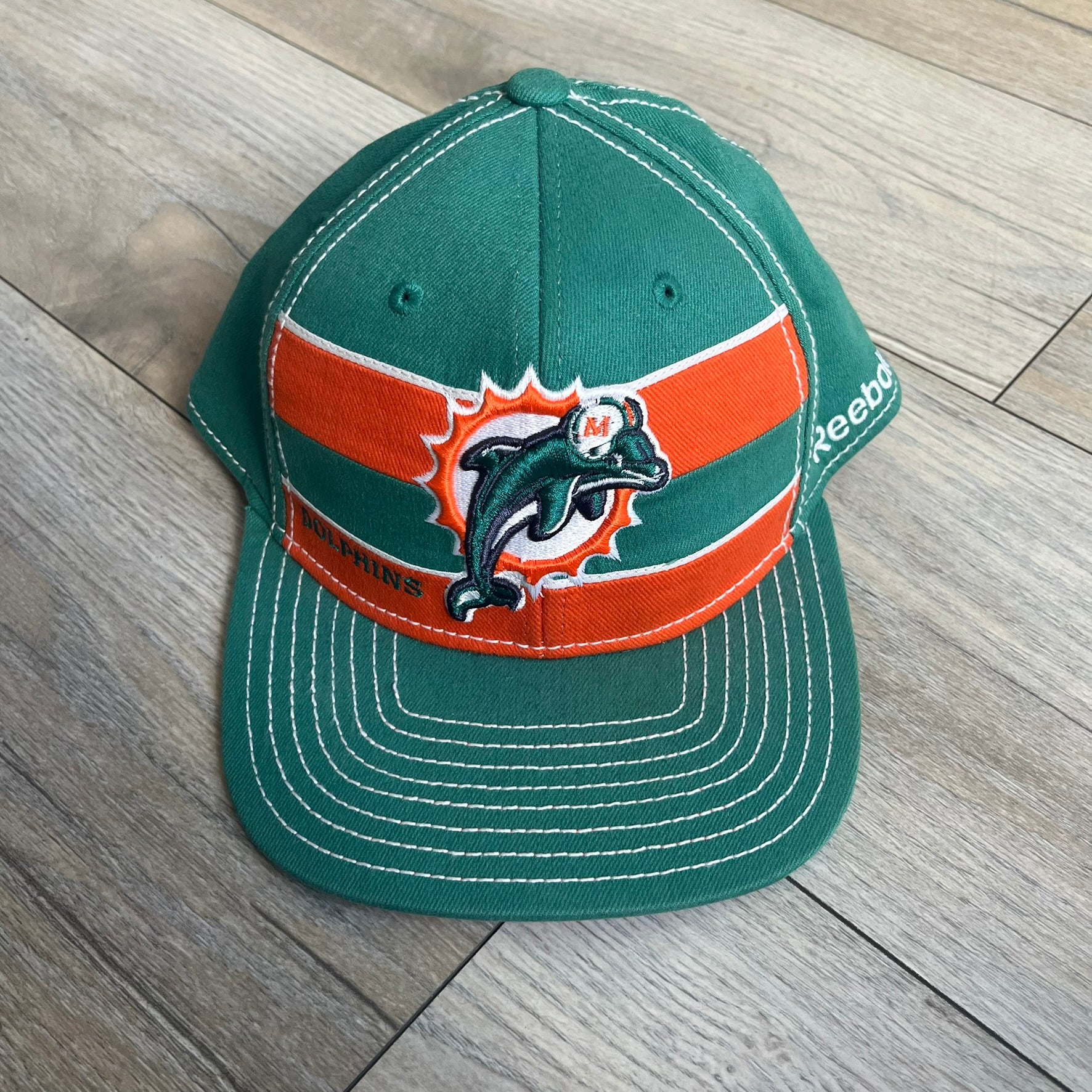 Reebok NFL Miami Dolphins Onfield Snapback Cap is a turquoise snapback featuring embroidered Miami Dolphins branding on the front, framed by two bold orange stripes positioned at the top and bottom of the front panel. The visor includes white stitching on the top surface and an orange underside for contrast. The side is detailed with a white Reebok logo, while the back features NFL Equipment and Onfield branding.