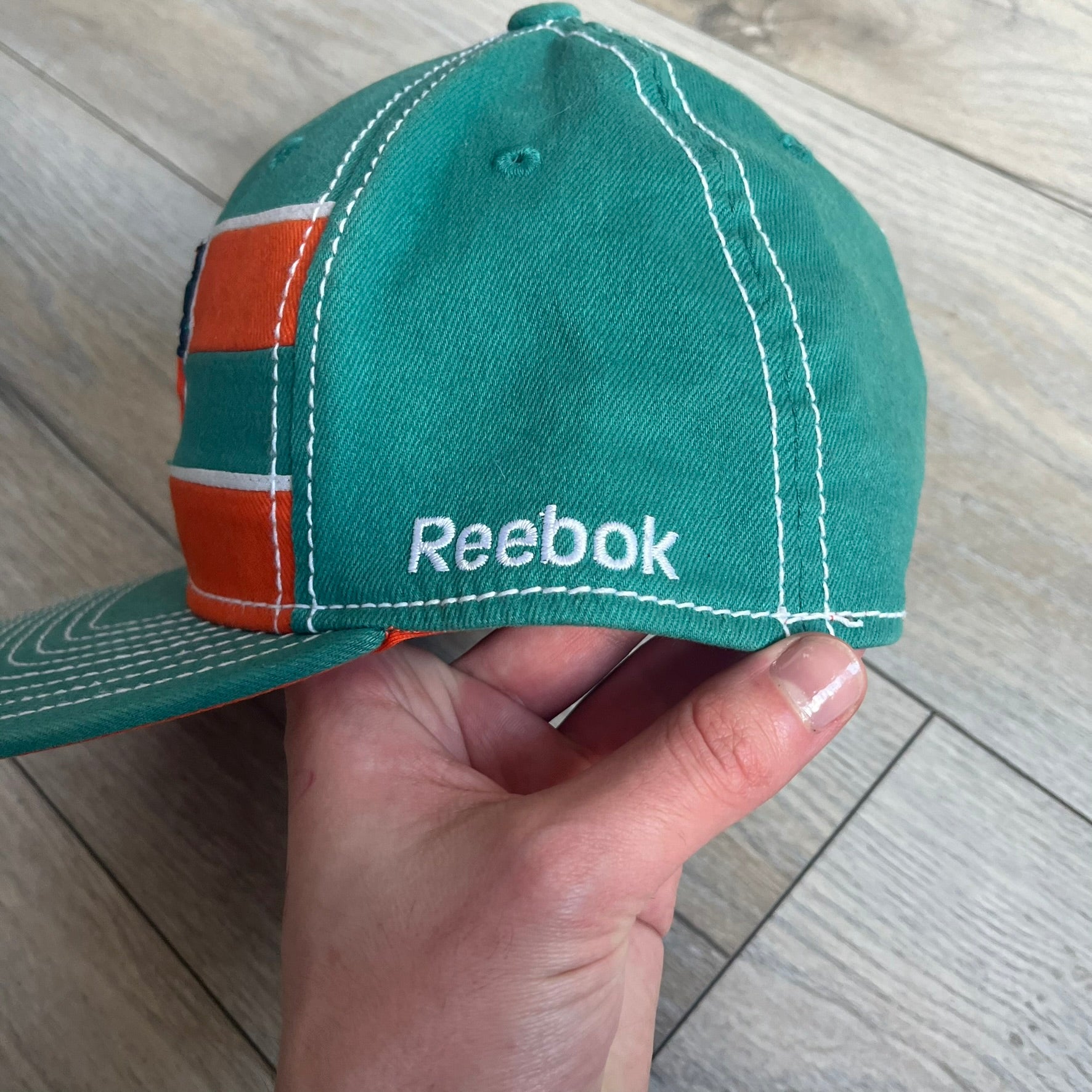 Reebok NFL Miami Dolphins Onfield Snapback Cap is a turquoise snapback featuring embroidered Miami Dolphins branding on the front, framed by two bold orange stripes positioned at the top and bottom of the front panel. The visor includes white stitching on the top surface and an orange underside for contrast. The side is detailed with a white Reebok logo, while the back features NFL Equipment and Onfield branding.