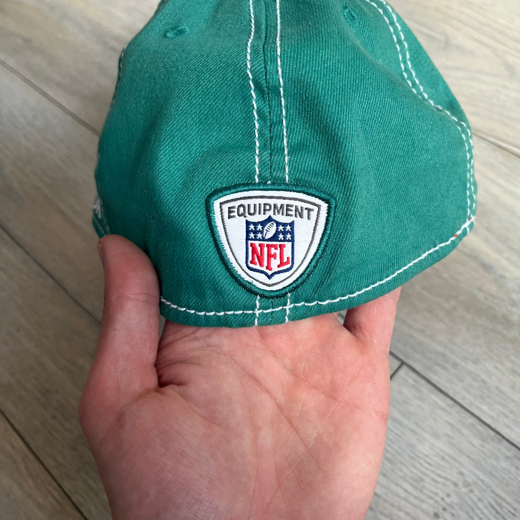 Reebok NFL Miami Dolphins Onfield Snapback Cap is a turquoise snapback featuring embroidered Miami Dolphins branding on the front, framed by two bold orange stripes positioned at the top and bottom of the front panel. The visor includes white stitching on the top surface and an orange underside for contrast. The side is detailed with a white Reebok logo, while the back features NFL Equipment and Onfield branding.