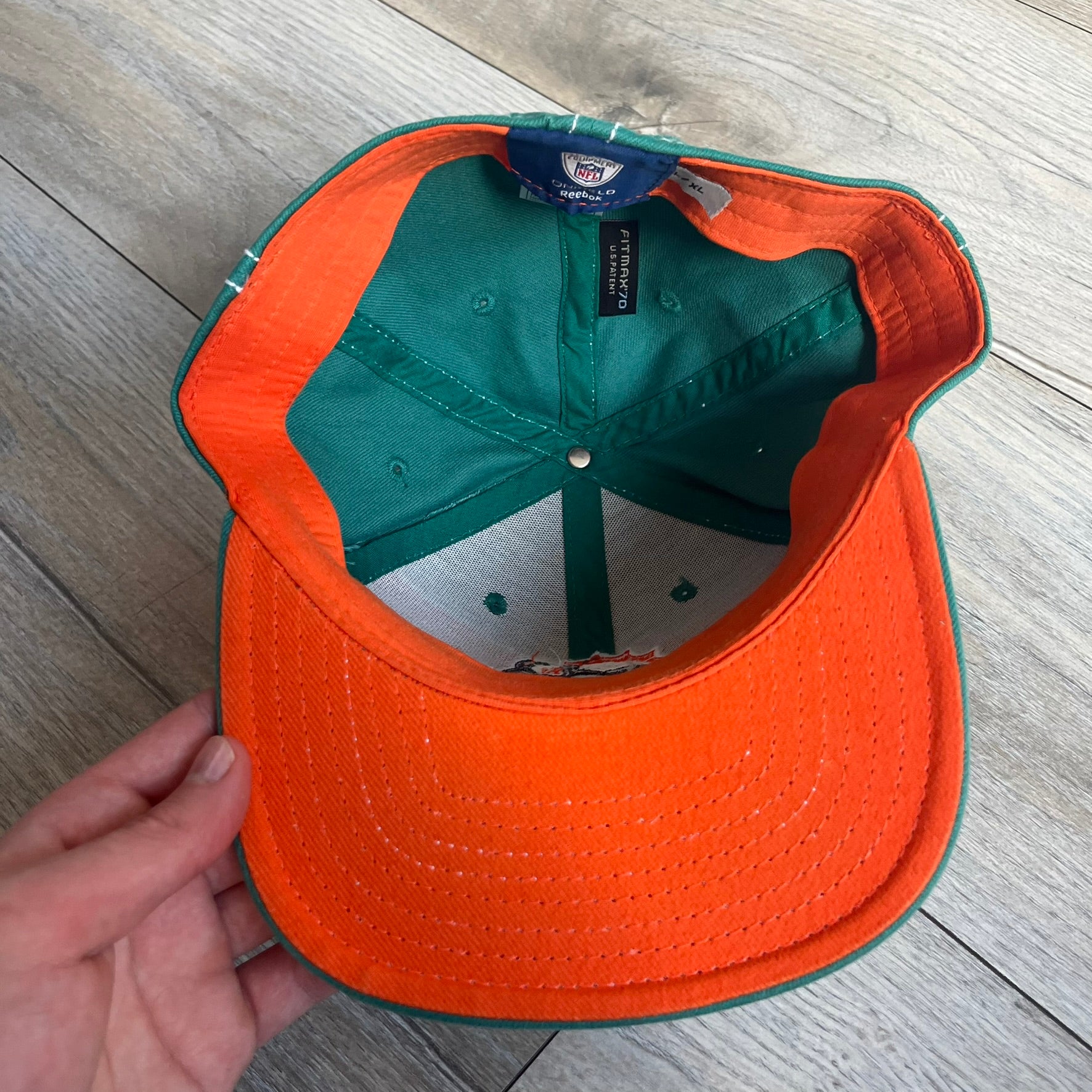 Reebok NFL Miami Dolphins Onfield Snapback Cap is a turquoise snapback featuring embroidered Miami Dolphins branding on the front, framed by two bold orange stripes positioned at the top and bottom of the front panel. The visor includes white stitching on the top surface and an orange underside for contrast. The side is detailed with a white Reebok logo, while the back features NFL Equipment and Onfield branding.