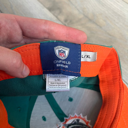 Reebok NFL Miami Dolphins Onfield Snapback Cap is a turquoise snapback featuring embroidered Miami Dolphins branding on the front, framed by two bold orange stripes positioned at the top and bottom of the front panel. The visor includes white stitching on the top surface and an orange underside for contrast. The side is detailed with a white Reebok logo, while the back features NFL Equipment and Onfield branding.