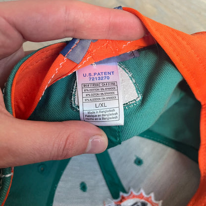 Reebok NFL Miami Dolphins Onfield Snapback Cap is a turquoise snapback featuring embroidered Miami Dolphins branding on the front, framed by two bold orange stripes positioned at the top and bottom of the front panel. The visor includes white stitching on the top surface and an orange underside for contrast. The side is detailed with a white Reebok logo, while the back features NFL Equipment and Onfield branding.