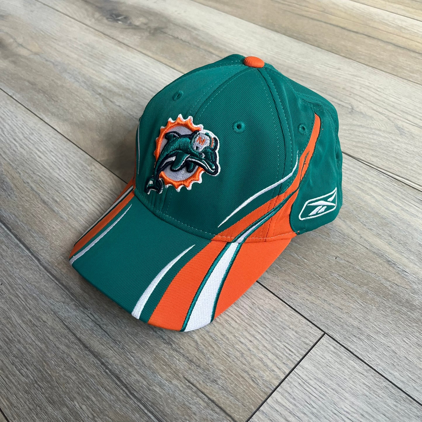 Reebok NFL Miami Dolphins Sideline Baseball Cap is a turquoise baseball cap featuring embroidered Miami Dolphins branding on the front. It includes orange and white accent detailing across the design. The side is finished with a white embroidered Reebok logo, while the back and inside features NFL Equipment tags. The construction uses cotton with a structured fit and a classic adjustable design.