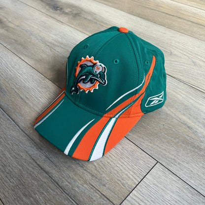 Reebok NFL Miami Dolphins Sideline Baseball Cap is a turquoise baseball cap featuring embroidered Miami Dolphins branding on the front. It includes orange and white accent detailing across the design. The side is finished with a white embroidered Reebok logo, while the back and inside features NFL Equipment tags. The construction uses cotton with a structured fit and a classic adjustable design.