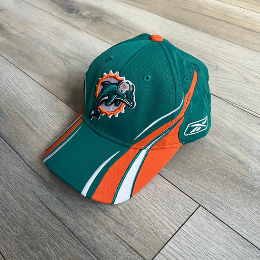 Reebok NFL Miami Dolphins Sideline Baseball Cap is a turquoise baseball cap featuring embroidered Miami Dolphins branding on the front. It includes orange and white accent detailing across the design. The side is finished with a white embroidered Reebok logo, while the back and inside features NFL Equipment tags. The construction uses cotton with a structured fit and a classic adjustable design.