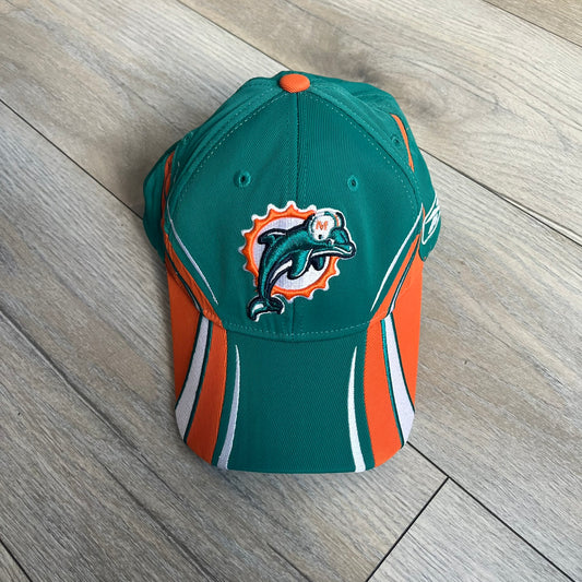 Reebok NFL Miami Dolphins Sideline Baseball Cap is a turquoise baseball cap featuring embroidered Miami Dolphins branding on the front. It includes orange and white accent detailing across the design. The side is finished with a white embroidered Reebok logo, while the back and inside features NFL Equipment tags. The construction uses cotton with a structured fit and a classic adjustable design.