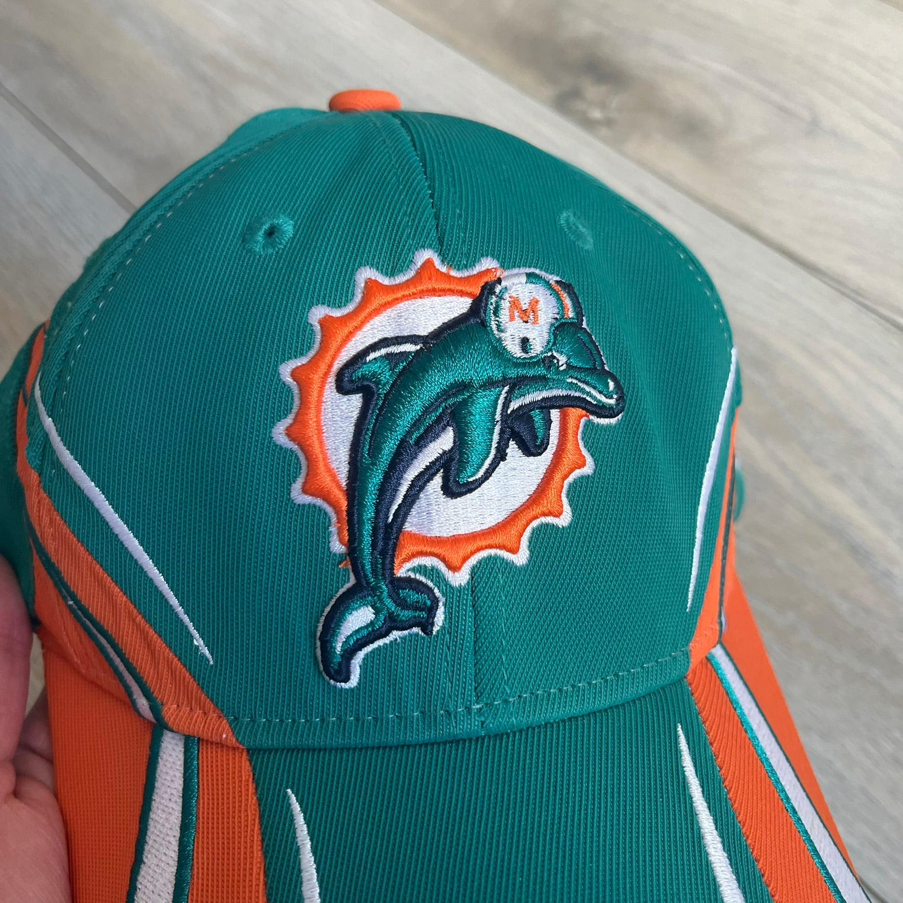Reebok NFL Miami Dolphins Sideline Baseball Cap is a turquoise baseball cap featuring embroidered Miami Dolphins branding on the front. It includes orange and white accent detailing across the design. The side is finished with a white embroidered Reebok logo, while the back and inside features NFL Equipment tags. The construction uses cotton with a structured fit and a classic adjustable design.